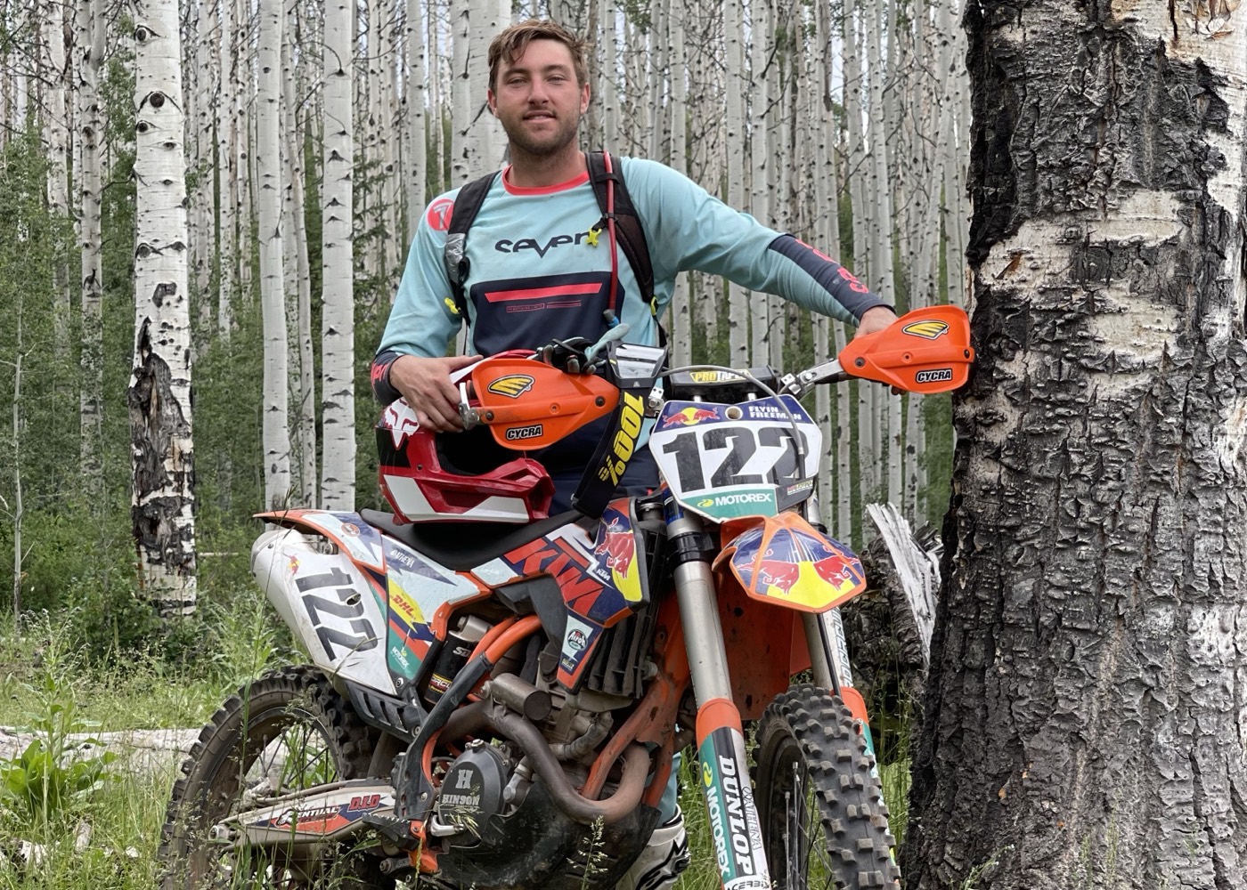 Dakota Freeman standing with his orange dirt bike in an aspen forest in the Colorado Rocky Mountains