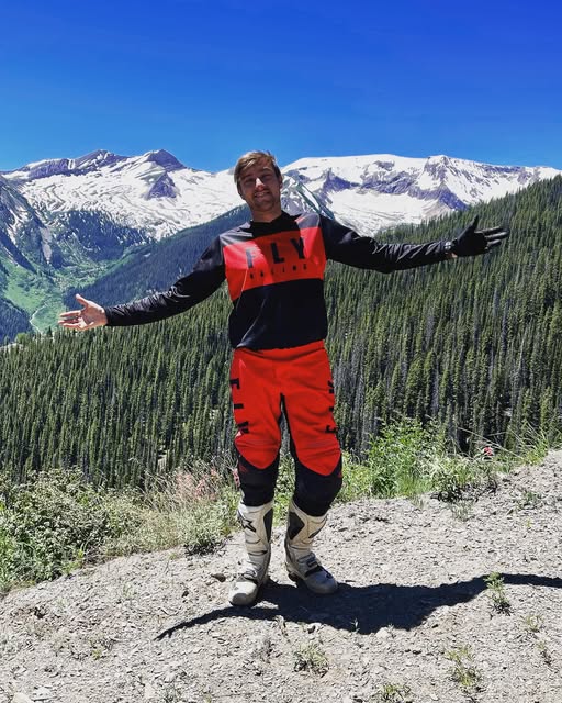 Dakota Freeman in red riding gear with arms open in front of snowy mountains